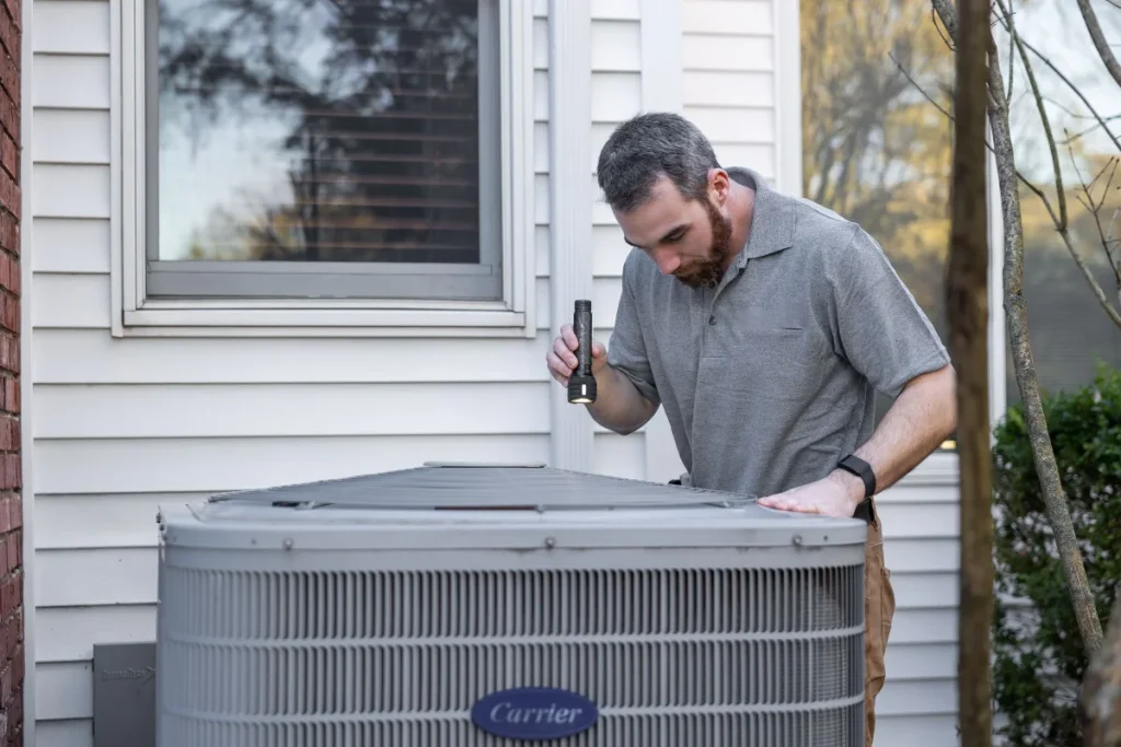 Technician checking a home condenser with a flashlight during a seasonal tune-up